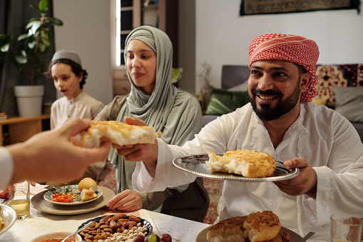 Selective focus shot of cheerful Muslim man sitting at table sharing flatbread during Uraza Bayram celebration dinner