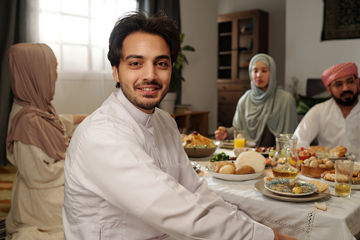 Portrait of happy young Muslim man wearing kandora sitting at festive table with his family on Eid Al-Fitr smiling at camera