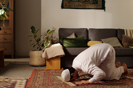 Young Middle Eastern man practicing namaz on prayer rug doing sujud in living room at home, copy space