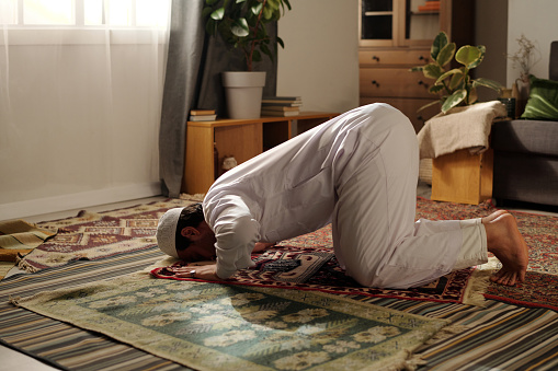 Young Muslim man wearing white clothes doing sajdah while practicing namaz on prayer rug in living room at home