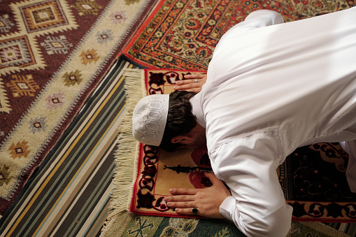 High angle view of unrecognizable young Muslim man wearing white clothes doing sadjah on prayer rug during namaz, copy space