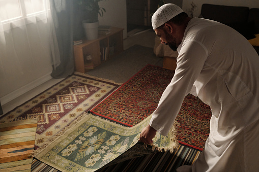 High angle view of Muslim man laying green prayer rug on floor in living room before doing namaz, copy space