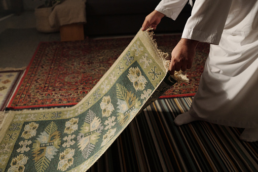 Unrecognizable Muslim man placing prayer rug on floor in living room at home before namaz, copy space