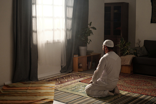 Wide shot of unrecognizable Muslim man sitting on prayer rug in living room at home praying salah during namaz, copy space