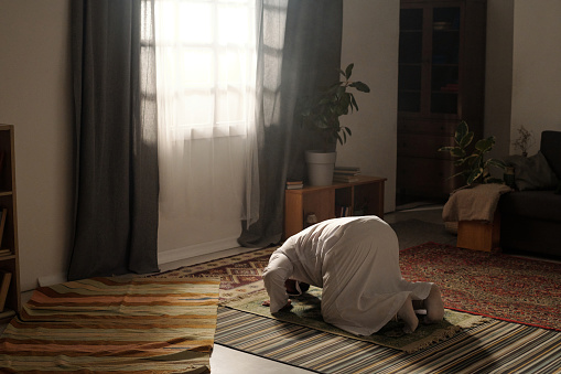Long shot of Muslim man wearing white thobe doing sujud on prayer rug during namaz in living room, copy space