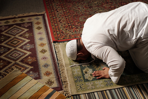 High angle view of mature Muslim man wearing white thobe and tuqiyah doing sajdah on prayer rug during namaz, copy space