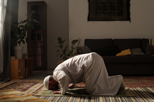 Long shot of mature bearded Muslim man wearing white thobe doing sajdah on prayer rug in living room at home, copy space