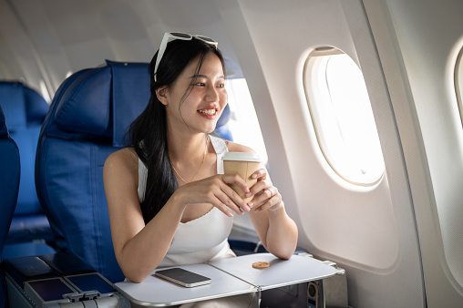 A happy, attractive Asian female passenger sipping coffee and enjoying the view outside the window during her flight, traveling by plane. people and transportation concepts