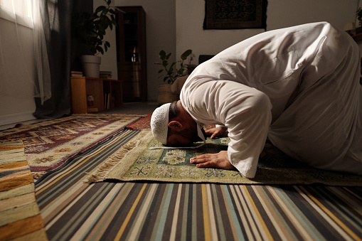 Side view of bearded Muslim man wearing white clothes praying salah on rug in living room doing sajdah, copy space