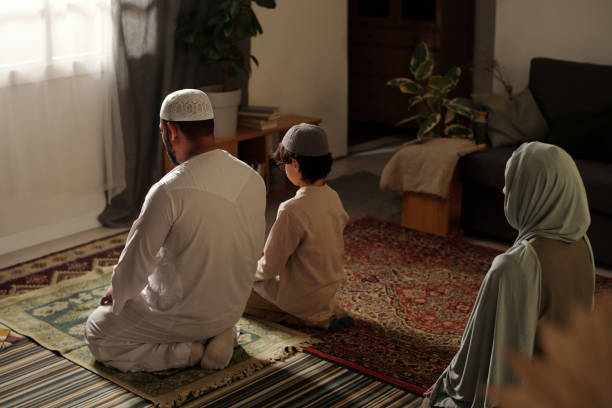 Modern Muslim Family Praying Salah High angle view of unrecognizable Muslim family with kid sitting on floor in living room praying salah father allah stock pictures, royalty-free photos & images