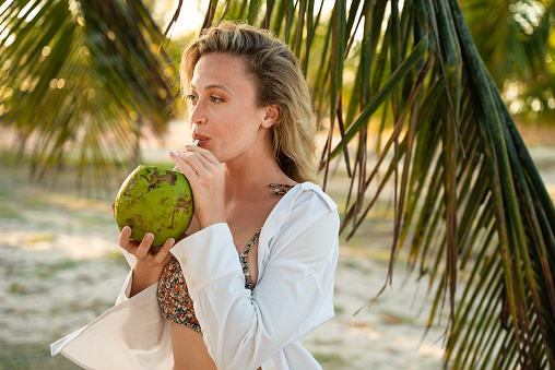 Charming woman on a brazilian beach drinking sweet water from coconut using a straw