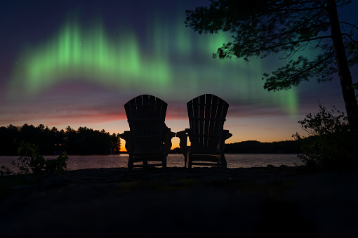 Two Adirondack chairs overlook a peaceful lake in Muskoka, Canada. It's nighttime at the cottage, with the chairs facing the colorful Aurora dancing in the sky.