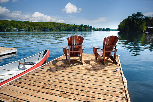 Tranquil Muskoka, Ontario setting as two Adirondack chairs grace a wooden dock by the shimmering waters of a calm lake on a sunlit summer day, accompanied by a canoe and paddles.