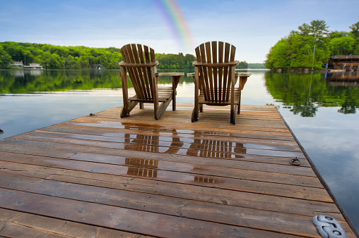 Two Adirondack chairs are positioned on a wet wooden pier, offering a tranquil view of a Muskoka lake. It's early morning at the cottage, with a vibrant rainbow adorning the sky.