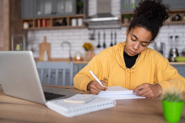 istock An african-american young woman is using laptop computer for remote work or studying online, she takes notes watching webinars or classes at kitchen table at home 2131819397