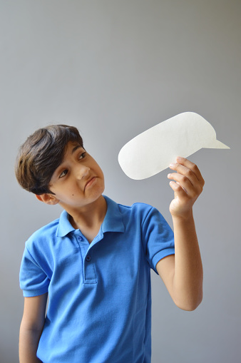 Vertical photograph of one 11 year old boy kid in casual blue colored polo t shirt holding a paper craft cut in shape of a blank empty speech bubble or call out and looking at it with strange astonished facial expressions with focus on hand and bubble over grey background