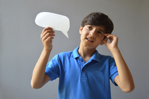 Horizontal photograph of one cute frowning 11 year old boy kid in casual blue colored polo t shirt holding a paper craft cut in shape of a blank empty speech bubble or call out thinking deeply with hand on forehead over grey background. Selective focus more on hand and bubble and soft focus on face.