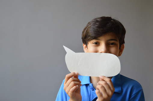 Horizontal photograph of one cute smiling  happy 11 year old boy kid in casual blue colored t shirt holding a paper craft cut in shape of a blank empty speech bubble or call out balloon as mask and looking with funny smile over gray background with copy space.