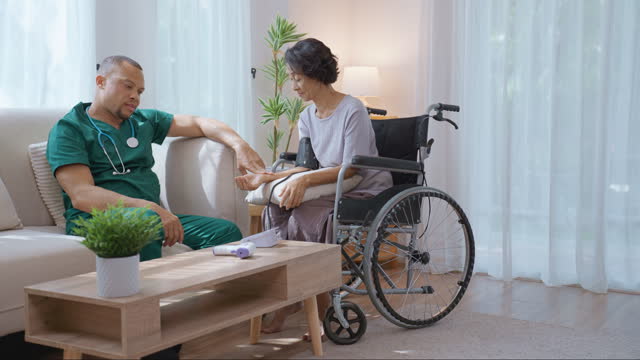 African male nurse providing home health consultation for a Thai senior woman patient who sitting in wheelchair and conducting a medical procedure by measuring the blood pressure for her medical check-up and illness prevention.