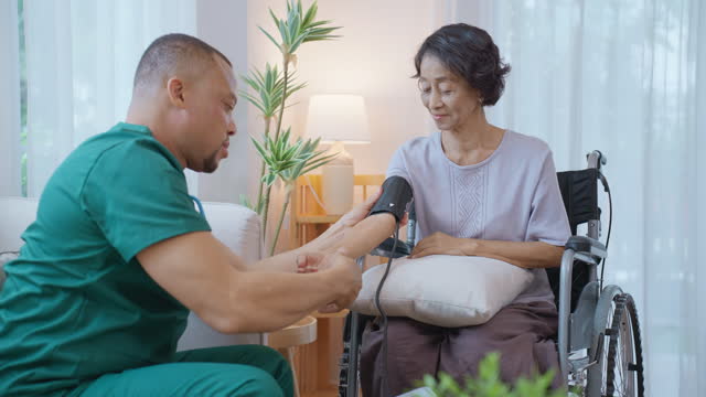 African male nurse conducting a medical procedure by measuring the blood pressure of a Thai senior woman who sitting in a wheelchair in her cozy living room for medical check-up and illness prevention.