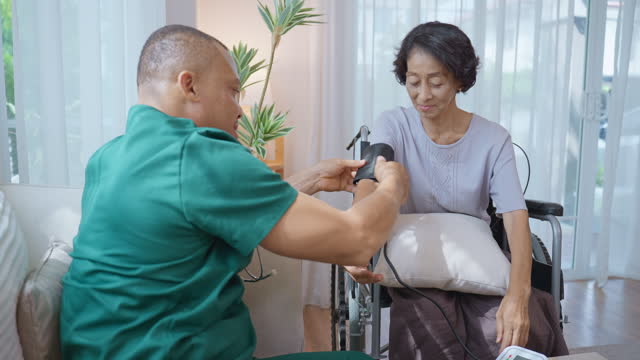 An African male nurse providing home healthcare services by measuring the blood pressure of a Thai senior woman patient who sitting in a wheelchair in her cozy living room for ensuring the woman's well-being.