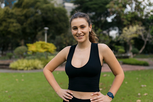 Portrait of a fit young woman in sportswear with hands on hips. Happy female model looking at camera and smiling at park, taking break from yoga workout.