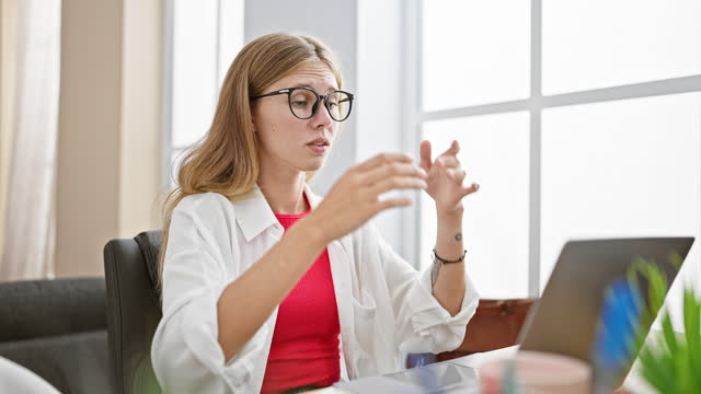 Stressed blonde woman in glasses feeling overwhelmed at an office desk with laptop, showing signs of workplace fatigue
