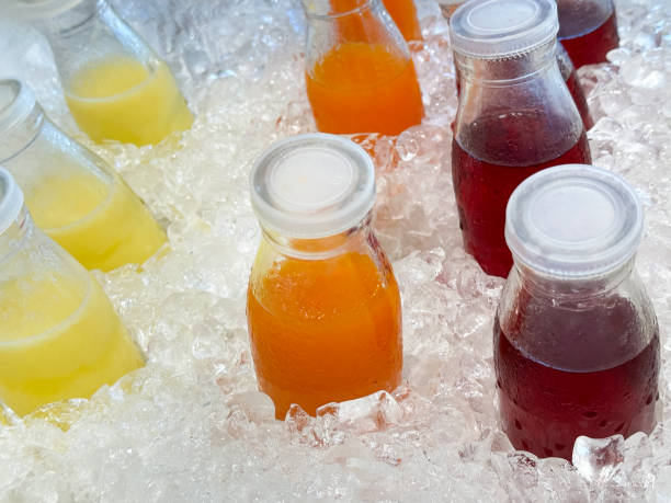 Full frame image of metal tray filled with chilled cold fruit juices drinks surrounded by ice cubes, crushed ice and screw top bottles of pineapple, orange and cranberry juices, non-alcoholic drinks in glass bottles covered in condensation, elevated view