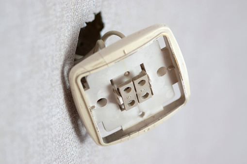 An exposed electrical power outlet missing its protective faceplate is visible against a white wall, revealing the internal components and wires, implying a potential safety hazard.
