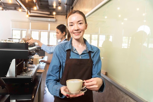 Happy woman waitress in apron standing near coffee machine holding cup with hot beverage