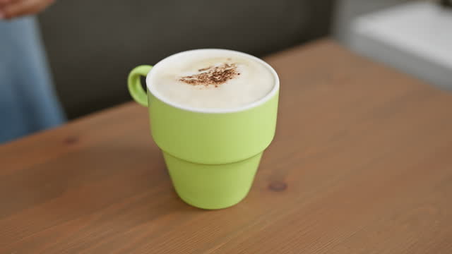 A hand gripping a green cup of cappuccino on a wooden table in an indoor setting