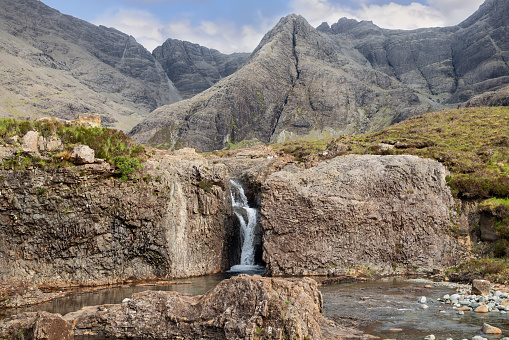 A solitary waterfall tumbles into the tranquil Fairy Pools, embraced by the craggy grandeur of the Cuillin Mountains on Scotland Isle of Skye