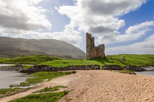 Ardvreck Castle, nestled against the Scottish Highlands, overlooks a tranquil lake with gentle waves caressing its shores. This landscape combines the rugged beauty of ancient ruins with the serene ambiance of a summer day in Scotland