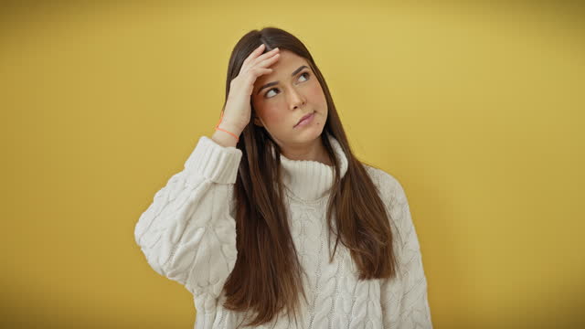 Worried young hispanic woman in sweater, hands over head, stressed with problems and crisis, standing over yellow isolated background