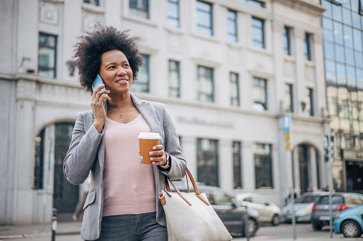 Smiling African woman walking down the street, talking on the Phone and holding cup of coffee. Coffee break. Building exterior in the background.