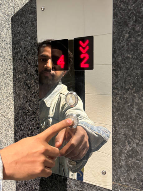Image of Indian man reflected in highly polished chrome surface of lift call button control panel, pushing down button with index finger, red LED floor number display and direction arrows, highly polished marble tiled wall, focus on foreground