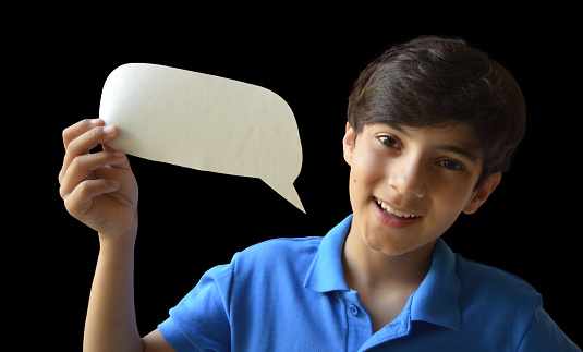 Horizontal photograph of cut out of one cute smiling happy 11 year old boy kid in casual blue colored polo t shirt holding a paper craft cut in shape of a blank empty speech bubble or call out pointing downwards to himself isolated over black background