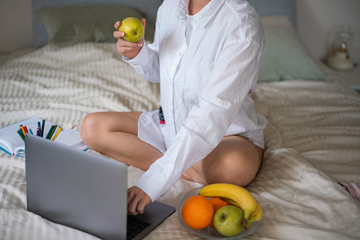 Unrecognizable woman holding apple in hand while working on laptop on bed in bedroom