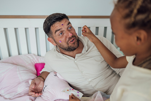 Over-the-shoulder shot of Step Daughter mischievously putting sticky gems on her Step-Fathers face. The girl is wearing casual clothing and the father is wearing a smart casual beige button-down shirt. Videos are available similar to this scenario.