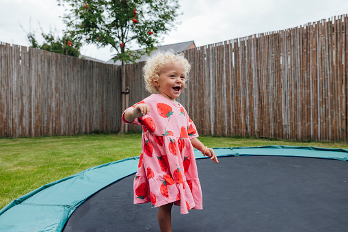 Medium shot of a female toddler in a sundress stood on an outdoor trampoline. The dress has cartoon strawberries on and she is smiling as she stands. Videos are available similar to this scenario.
