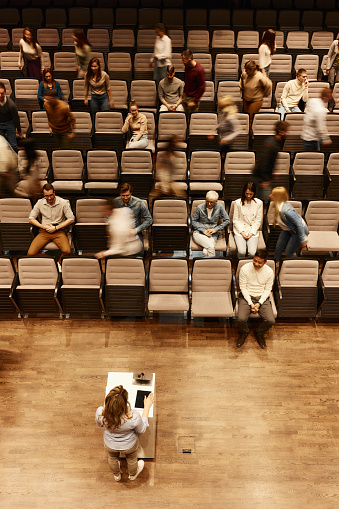 High angle view of crowd of people listening to a public speaker on a seminar at convention center while some of them are leaving in blurred motion. Copy space.