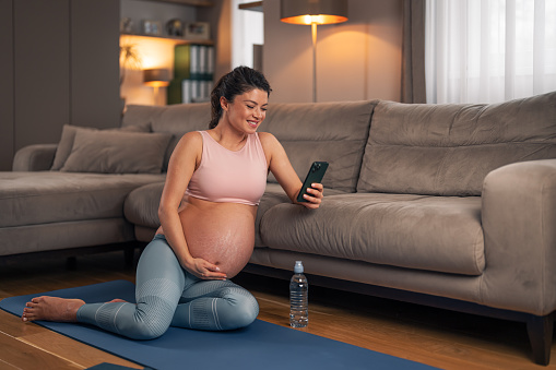 A happy pregnant lady holding her round belly and using her phone while taking a break from a home workout routine and leaning on a couch in the living room.