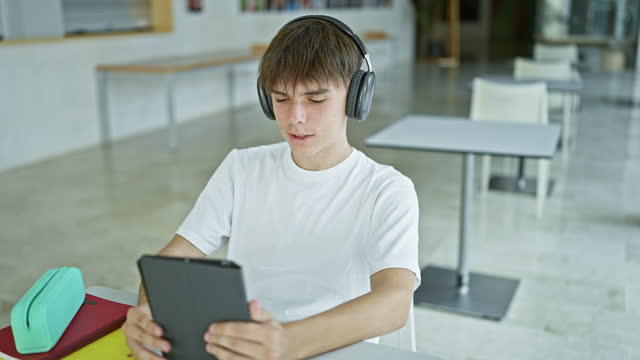 Young caucasian male student with headphones using tablet in modern university library