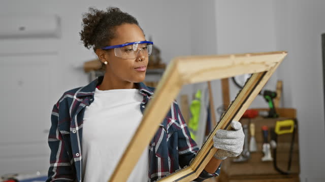 Focused woman with protective eyewear examines wooden frame in a well-equipped carpentry workshop