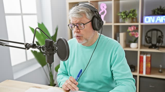 Grey-haired man in headphones speaks into microphone in radio studio, pen in hand, on air sign illuminating.