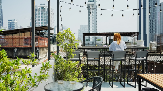 Rear view of young woman sitting at rooftop cafe table working on laptop