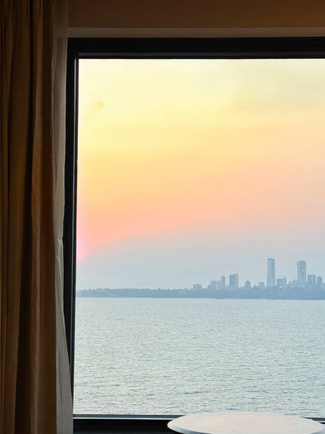 Stock photo showing close-up view of a table positioned in front of a picture window for view over cityscape skyscraper skyline view of Mumbai, Maharashtra, India at sunset. imagen de la vista de la puesta del sol del mar y del paisaje urbano de mumbai, maharashtra, india vista a través de un ventanal, mesa de la habitación doméstica, cortinas, alfombra, atmosférica, cielo de la puesta del sol naranja cambiante, enfoque en