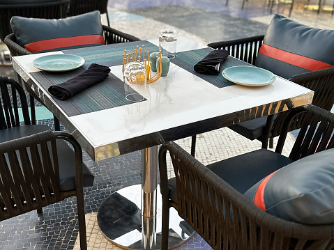 Close-up image of marble topped square tables and rattan wickerwork chairs with synthetic leather cushions on rooftop restaurant balcony, overlooking city scape, skyline and sea view on sunny afternoon, Mumbai, Maharashtra, India