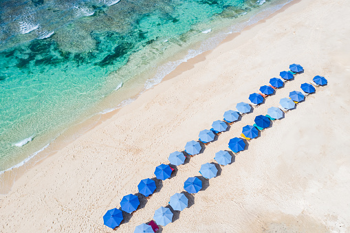 Bali - Melasti Beach with clear turquoise waters meeting the sandy shore where colorful beach umbrellas  are arranged in rows, captured from an aerial perspective.