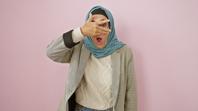 Middle-aged hispanic woman in hijab covers face with a hand, mirroring pure shock. peeking through fingers, she unveils an embarrassed expression over isolated pink background.